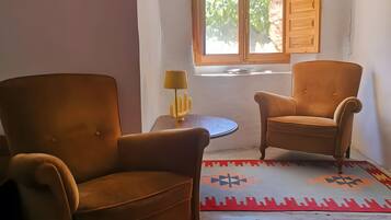 Quarto com duas poltronas marrons, uma mesinha de centro com abajur, um tapete estampado e uma janela com persianas de madeira.