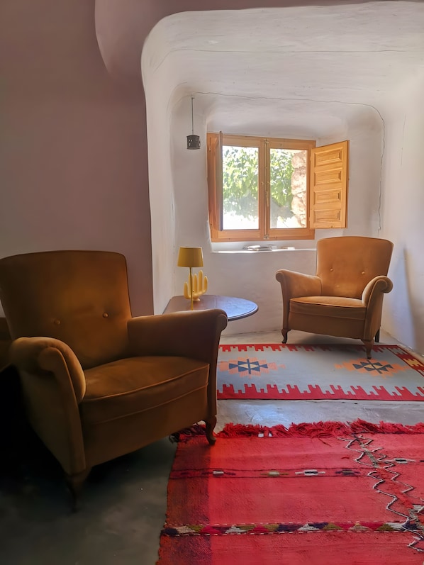 A room with two brown armchairs, a small table with a lamp, a patterned rug, and a window with wooden shutters. - Charming Cave in Andalusia with Private Terrace and Garden (Fuente Nueva)