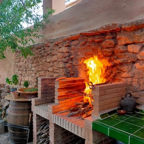 An outdoor brick oven with a fire burning inside, surrounded by potted plants and a metal grill. - Charming Cave in Andalusia with Private Terrace and Garden (Fuente Nueva)