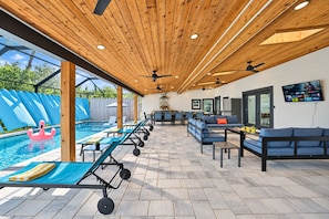 An outdoor pool area with lounge chairs, a barbecue setup, and a TV screen mounted on the wall. - Casa Lumina | 6 Bedroom Luxury Estate | Pool, Spa & Outdoor Kitchen (Temple Terrace)