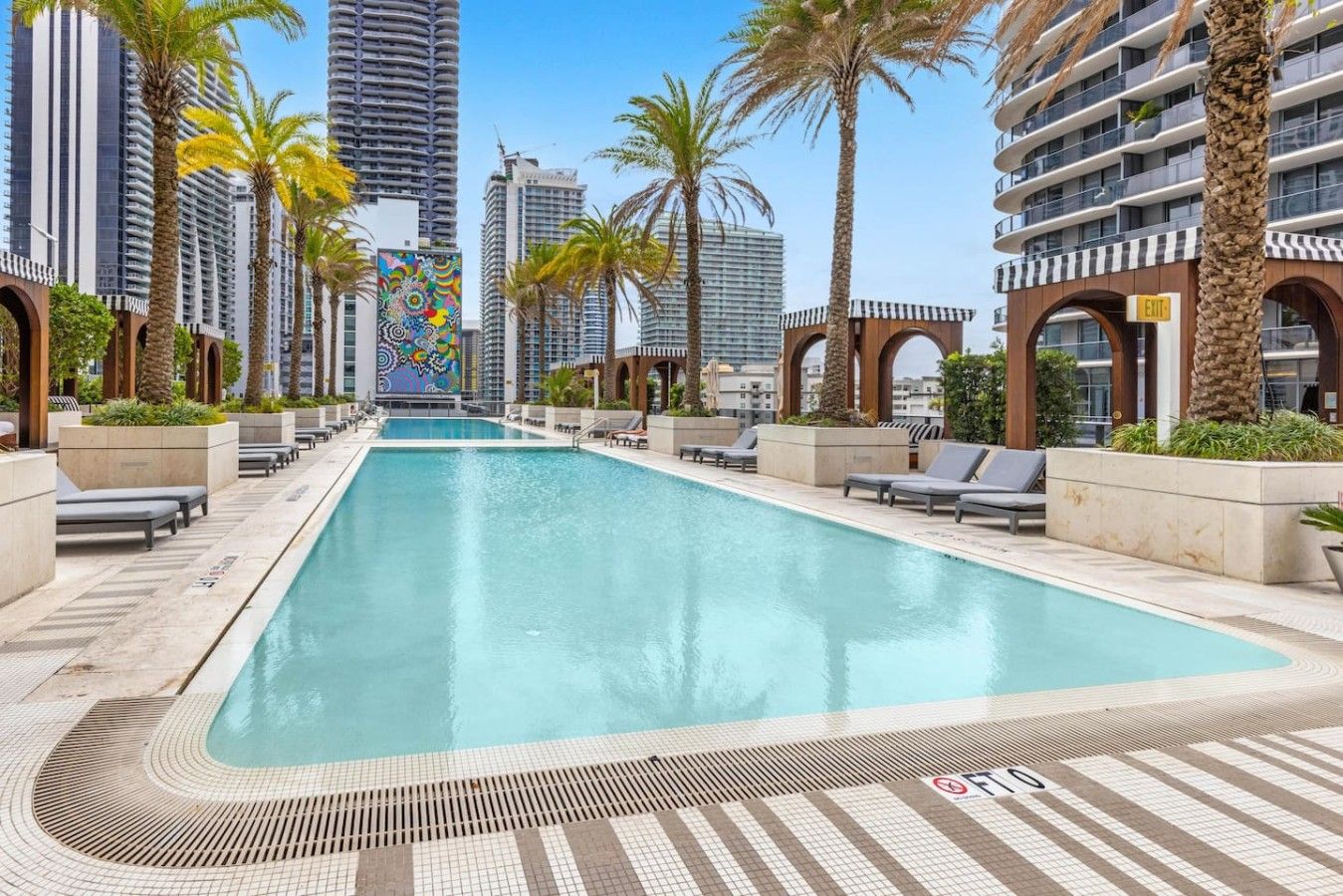 Una piscina con sillas de descanso, palmeras y edificios altos al fondo.