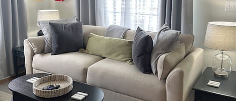 A living room with a sofa, a coffee table, a lamp, a guitar, and a window with curtains.