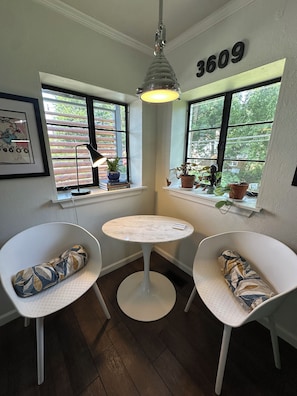 A corner room with a round table, two white chairs, a pendant light, and a window with a number sign.