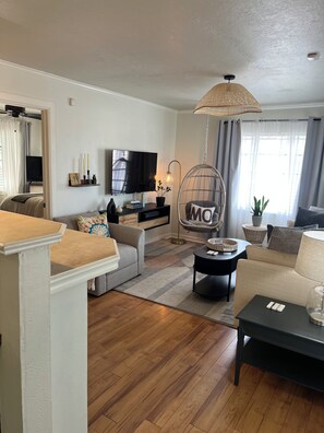 Un salon comprenant un canapé, une table basse, un téléviseur, un fauteuil suspendu et une grande fenêtre avec des rideaux.