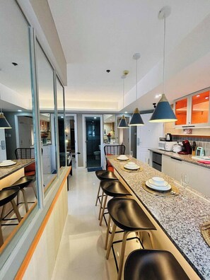 A modern kitchen with a long countertop, bar stools, pendant lighting, and a view into the dining area. - 2BR Tropical Scandi Suite (Paranaque)