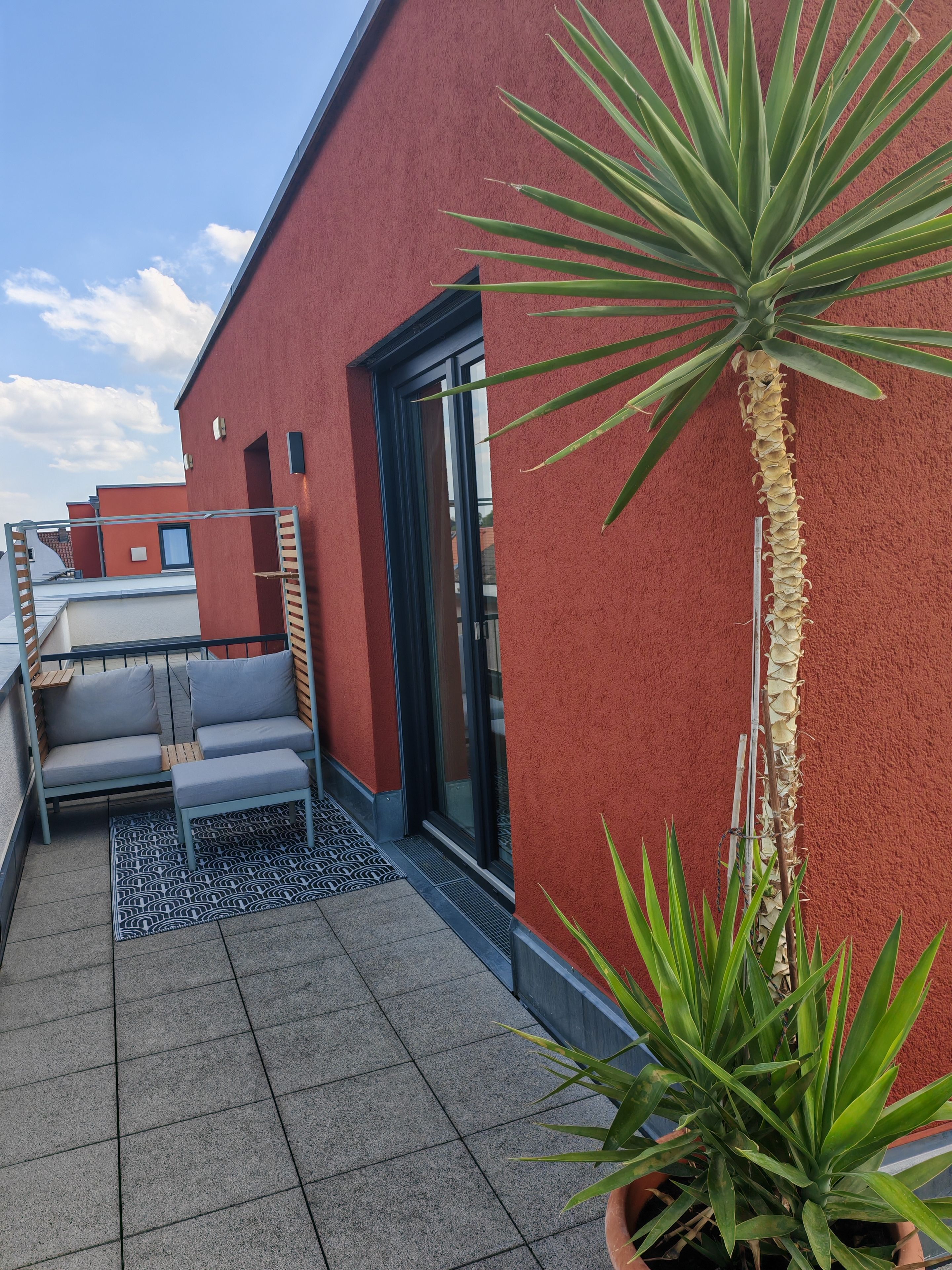 Une terrasse sur le toit avec un mur rouge, une porte-fenêtre coulissante, un petit coin salon avec des coussins et des plantes en pot.