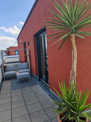 Una terraza en la azotea con un muro rojo, puerta corredera de vidrio, un rincón de estar con cojines y plantas en macetas.
