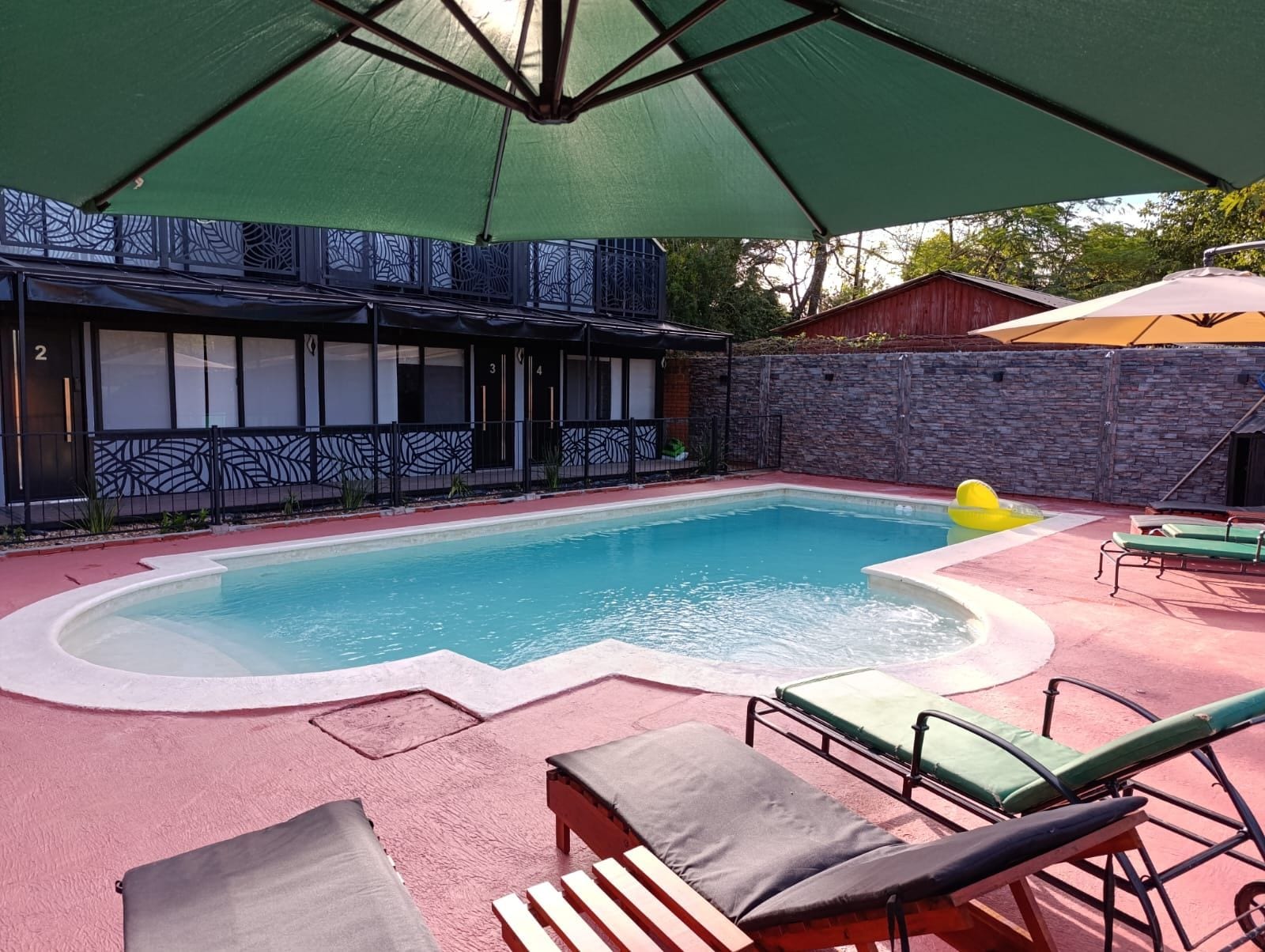 Una piscina con sillas de descanso y sombrillas, rodeada por un muro de piedra y un edificio con ventanas con diseños.