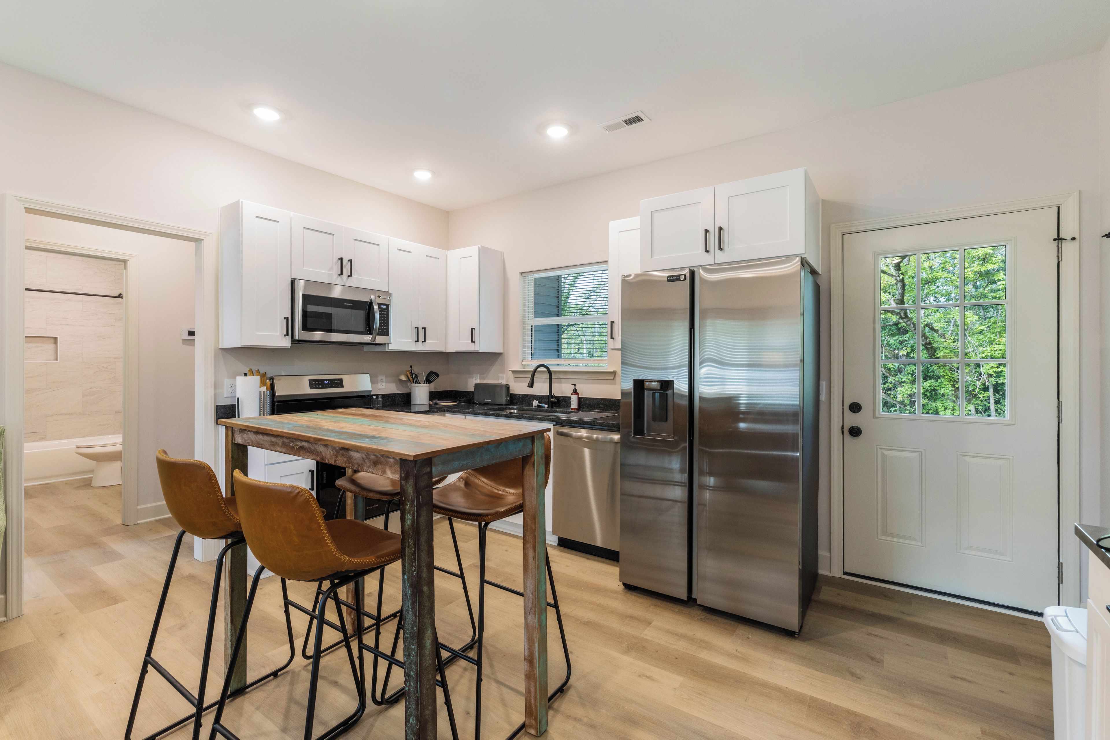 A modern kitchen with white cabinets, stainless steel appliances, a wooden table with bar stools, and a view of greenery through a window.