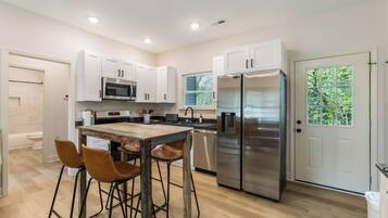 A modern kitchen with white cabinets, stainless steel appliances, a wooden table with bar stools, and a view of greenery through a window.