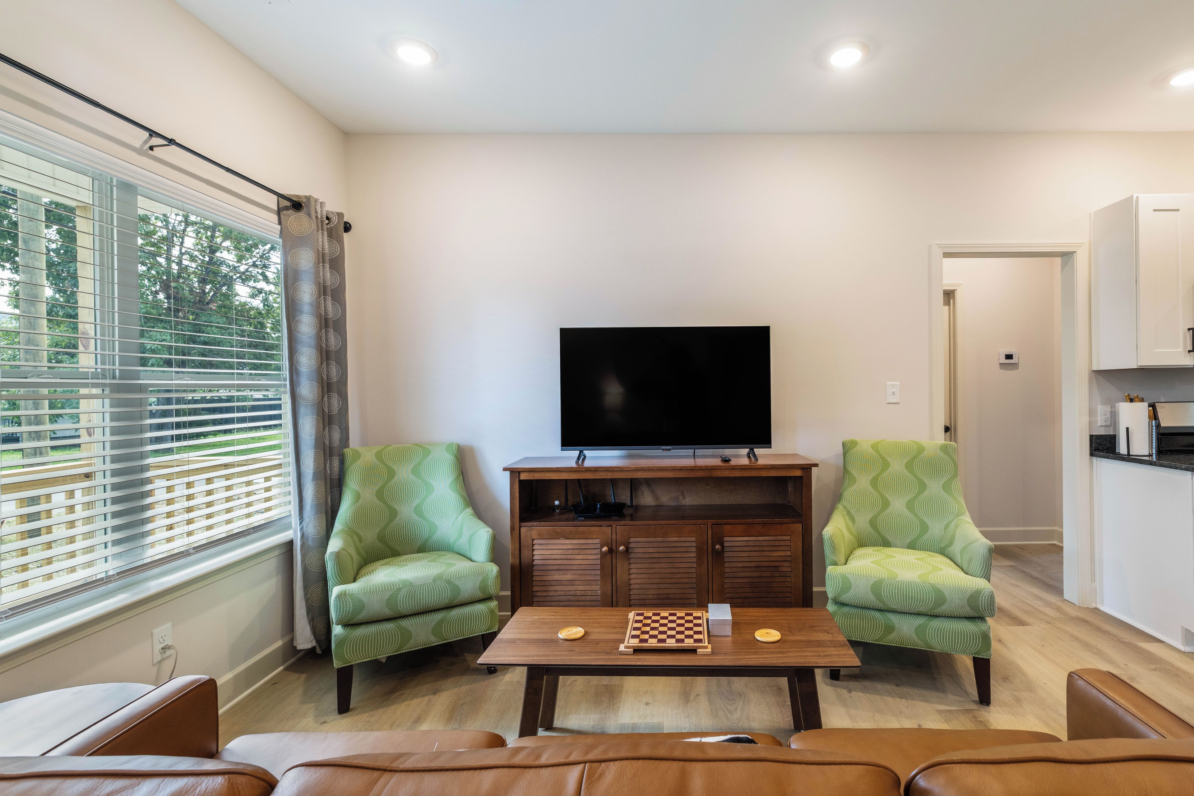 A living room with a flat-screen TV, two patterned armchairs, a coffee table with a chessboard, and a window with blinds.