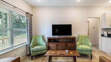 A living room with a flat-screen TV, two patterned armchairs, a coffee table with a chessboard, and a window with blinds.