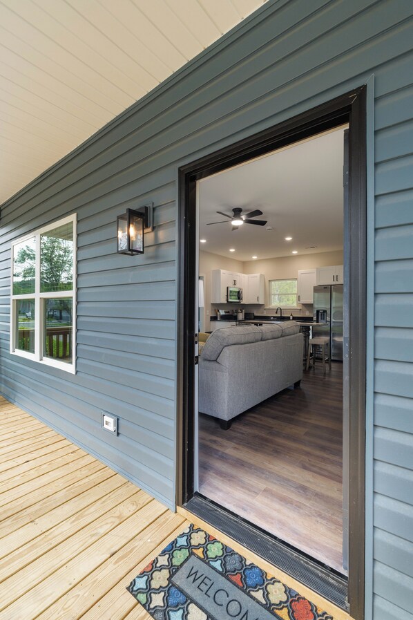 Une entrée résidentielle avec une terrasse en bois, un paillasson et une vue sur un espace de vie comprenant un canapé et des appareils de cuisine.