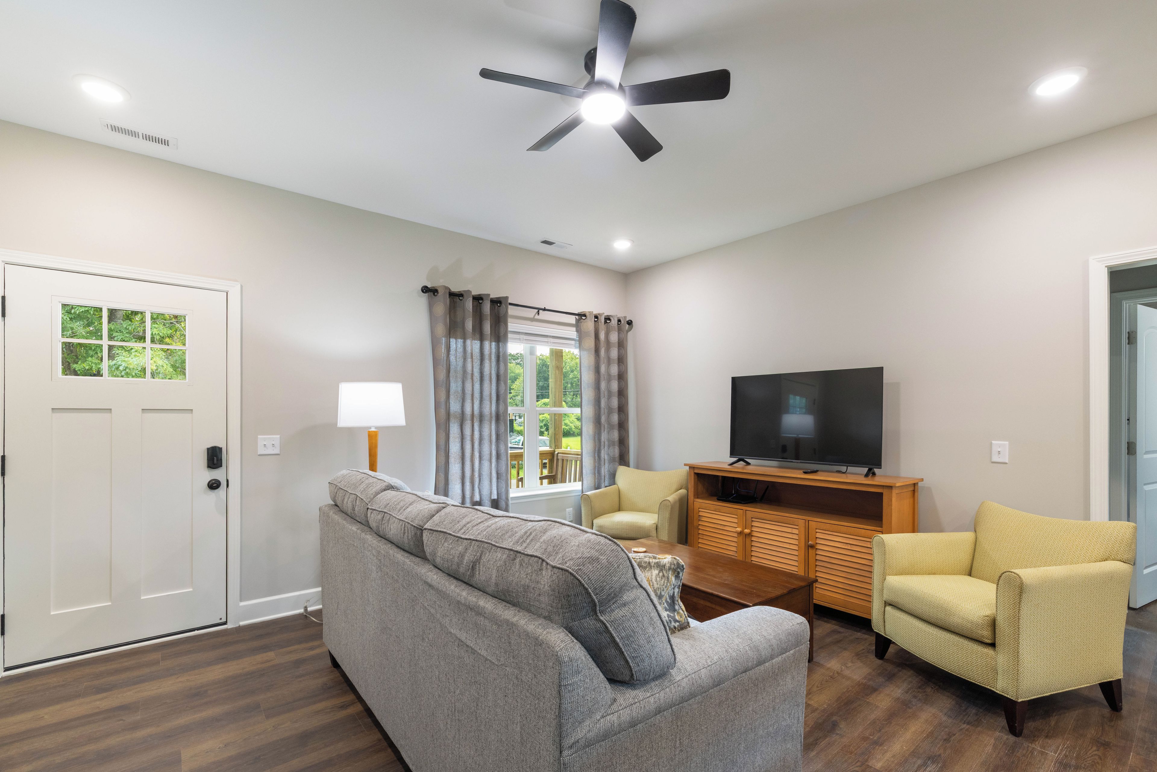 A living room with a ceiling fan, a television on a wooden stand, a grey sofa, a yellow armchair, and a window with curtains.