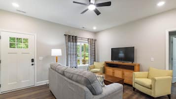 A living room with a ceiling fan, a television on a wooden stand, a grey sofa, a yellow armchair, and a window with curtains.