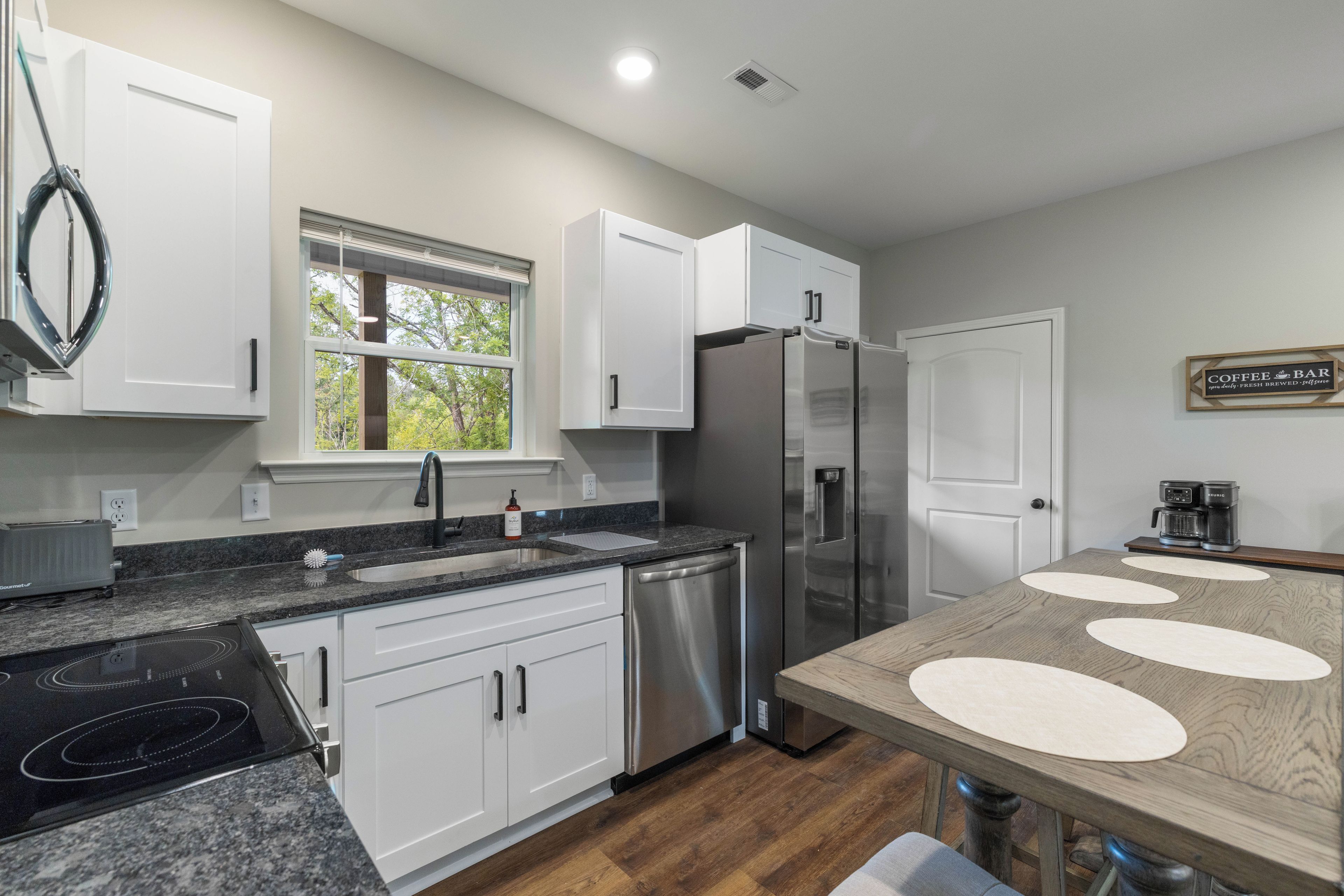A modern kitchen with white cabinets, a stainless steel refrigerator, a coffee bar sign, and a window with a view of trees.