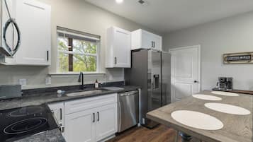 A modern kitchen with white cabinets, a stainless steel refrigerator, a coffee bar sign, and a window with a view of trees.