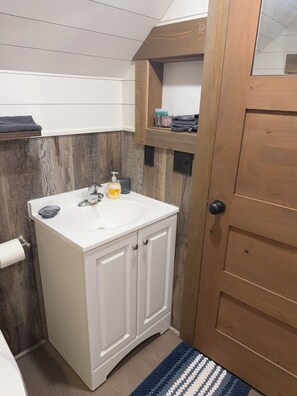A compact bathroom with a white sink, wooden cabinets, a towel rack, and a door with a handle. - #3 Glamping Wagon (Lake of the Woods)