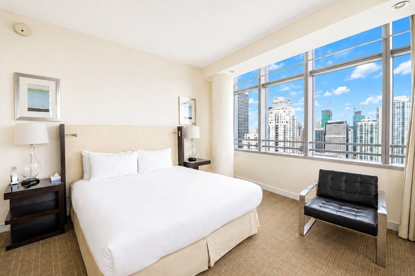 A hotel room with a large bed, two bedside tables, a chair, and a large window with a city view.