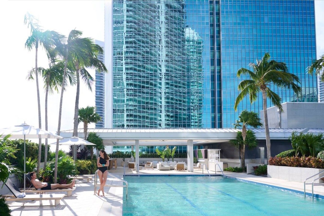 A pool area with lounge chairs and umbrellas, surrounded by palm trees and a modern building with reflective glass windows.