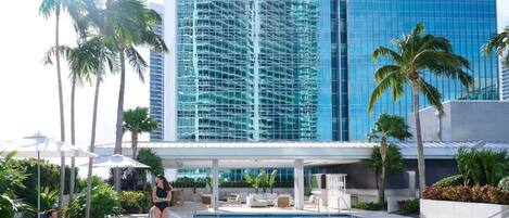 A pool area with lounge chairs and umbrellas, surrounded by palm trees and a modern building with reflective glass windows.
