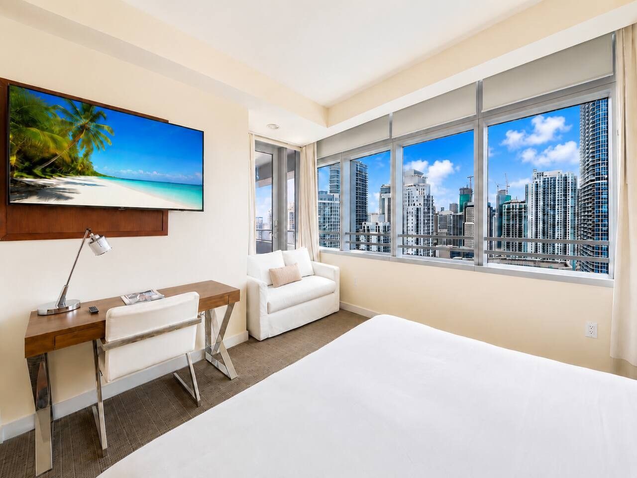 A hotel room with a large bed, a desk, a white sofa, and a city view through large windows.
