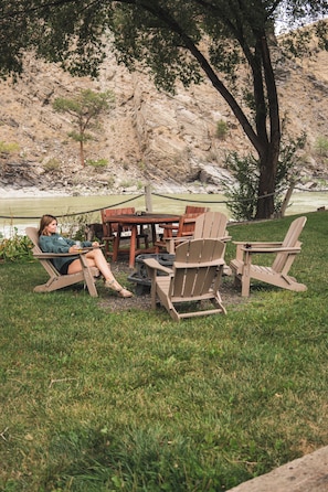 A person relaxing on a lounge chair by a wooden table and chairs, with a grassy area and a rocky hillside in the background.