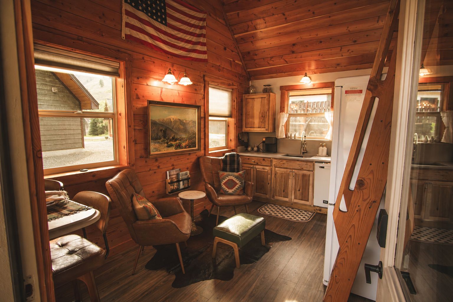 A cozy cabin interior with wooden walls, a dining area with a table and chairs, a kitchen with a sink and cabinets, and a window with an American flag.