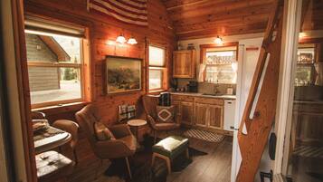 A cozy cabin interior with wooden walls, a dining area with a table and chairs, a kitchen with a sink and cabinets, and a window with an American flag.