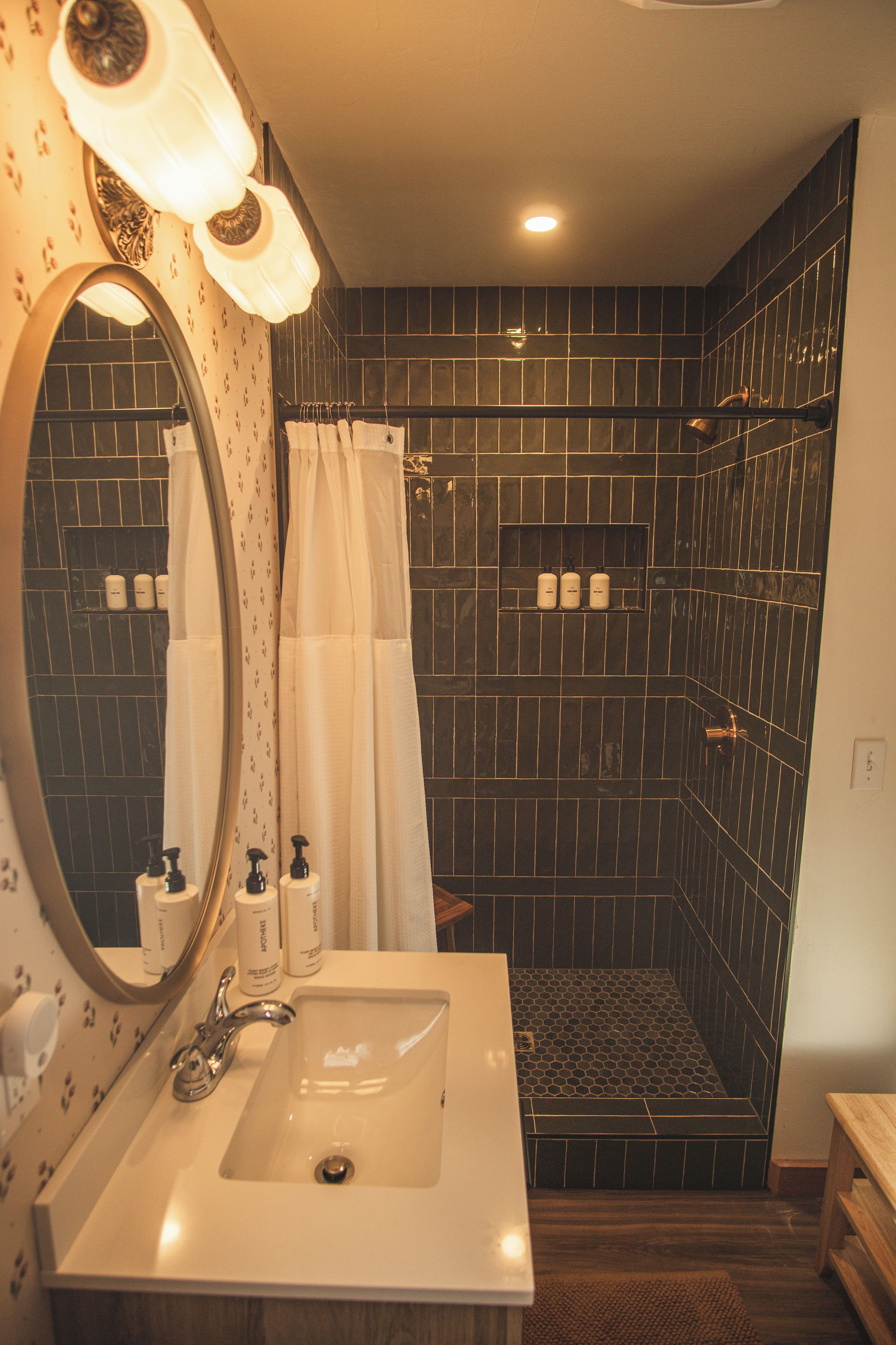 Un baño con lavamanos blanco, espejo, ducha con azulejos oscuros y artículos de aseo en la pared.