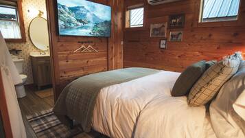 A wooden cabin bedroom with a bed, a mounted TV, a window, and a bathroom.
