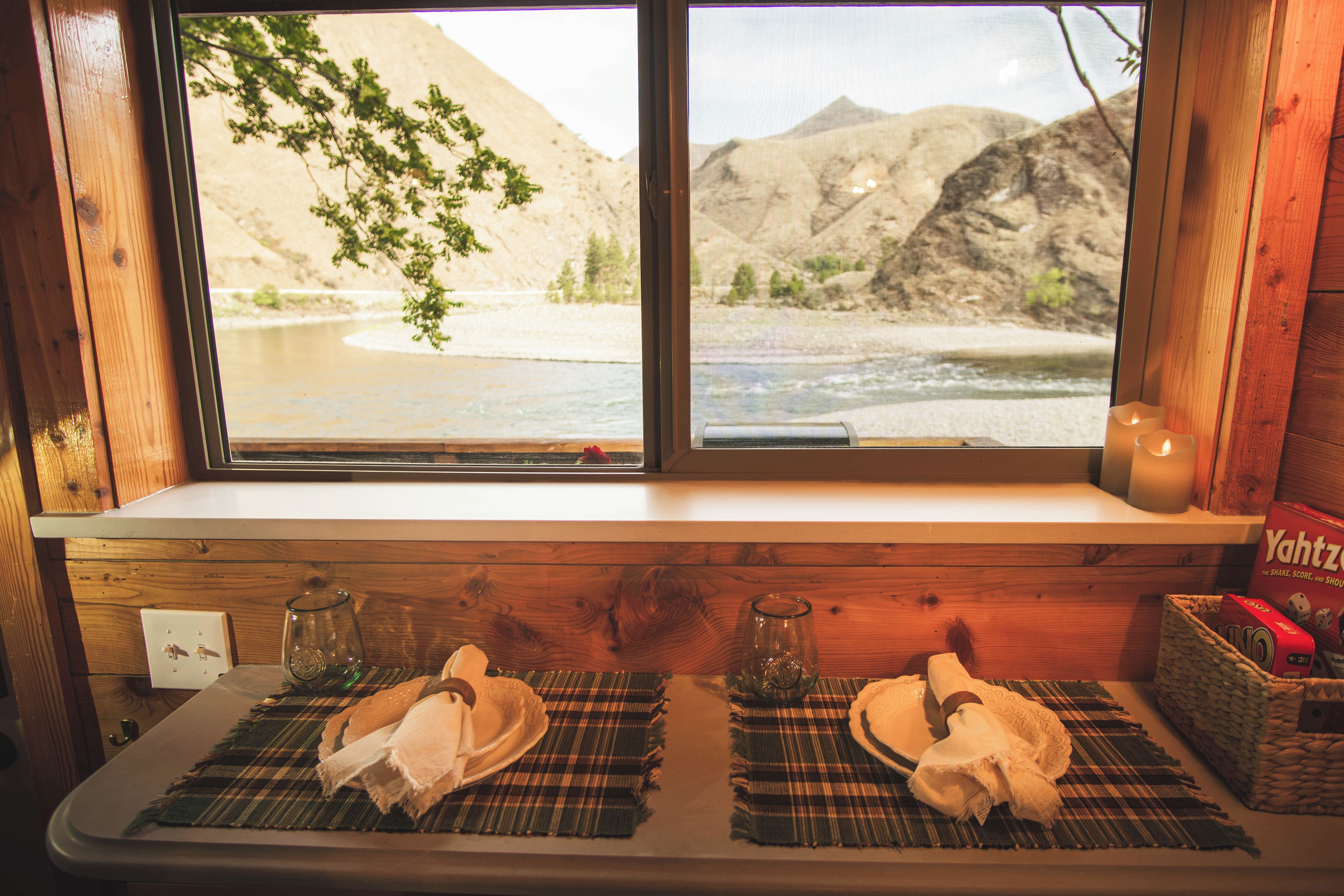 Janela com vista para um rio e montanhas, uma mesa com toalha de lugar, uma vela e um conjunto de dados.