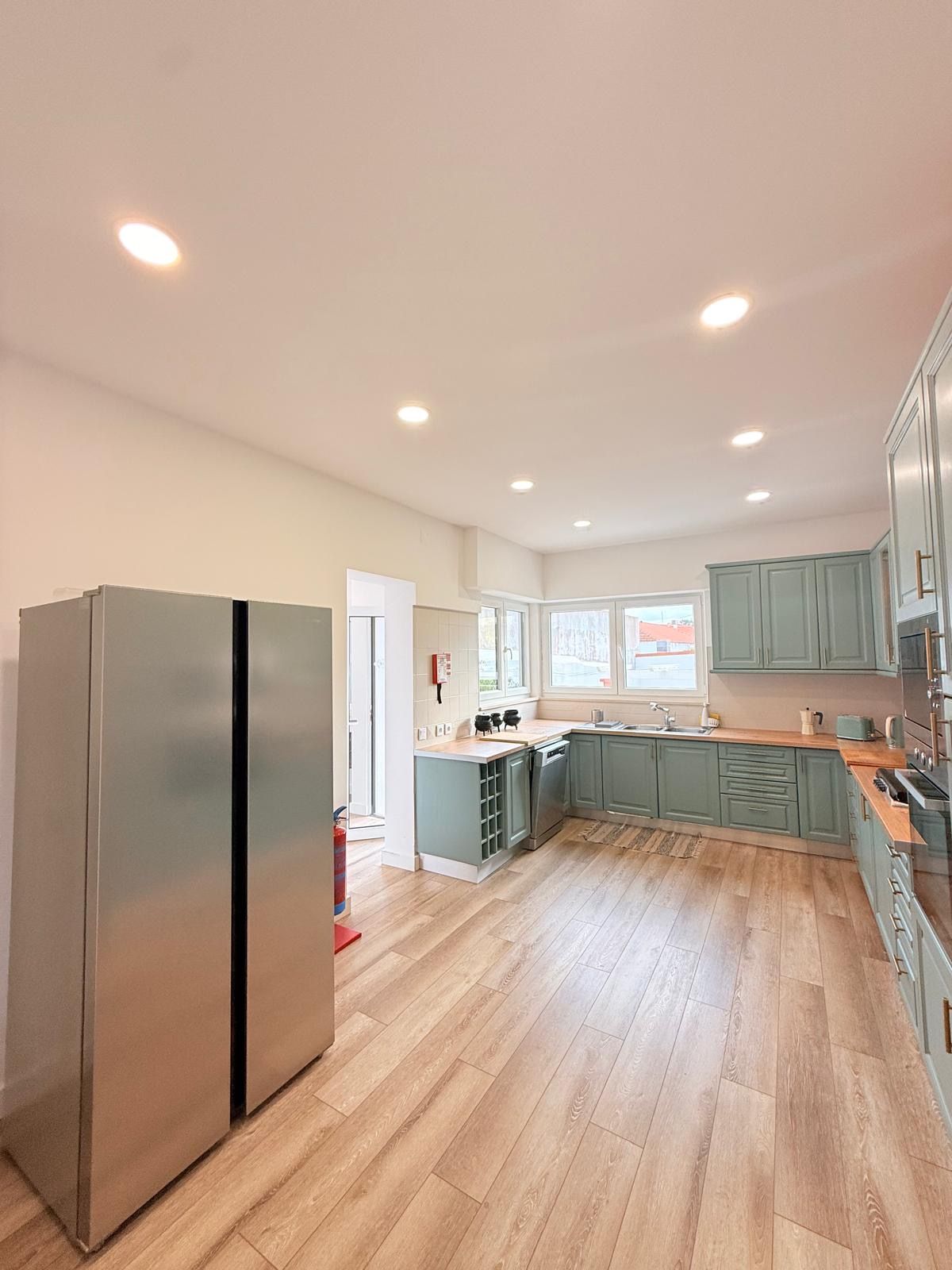 A spacious kitchen with a large refrigerator, wooden flooring, and light green cabinets.