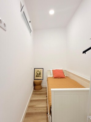 Ein enger Flur mit einem Bett auf der rechten Seite, einem kleinen Korbwäscheschrank und einem gerahmten Bild an der Wand.