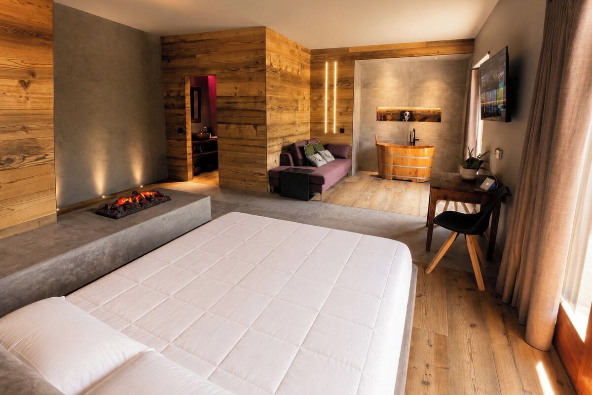 Quarto de hotel moderno com uma cama grande, uma parede de destaque em madeira, um pequeno espaço de estar e uma televisão.