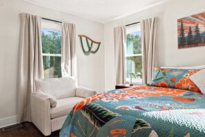 Un dormitorio con una cama, una silla y dos ventanas con cortinas.
