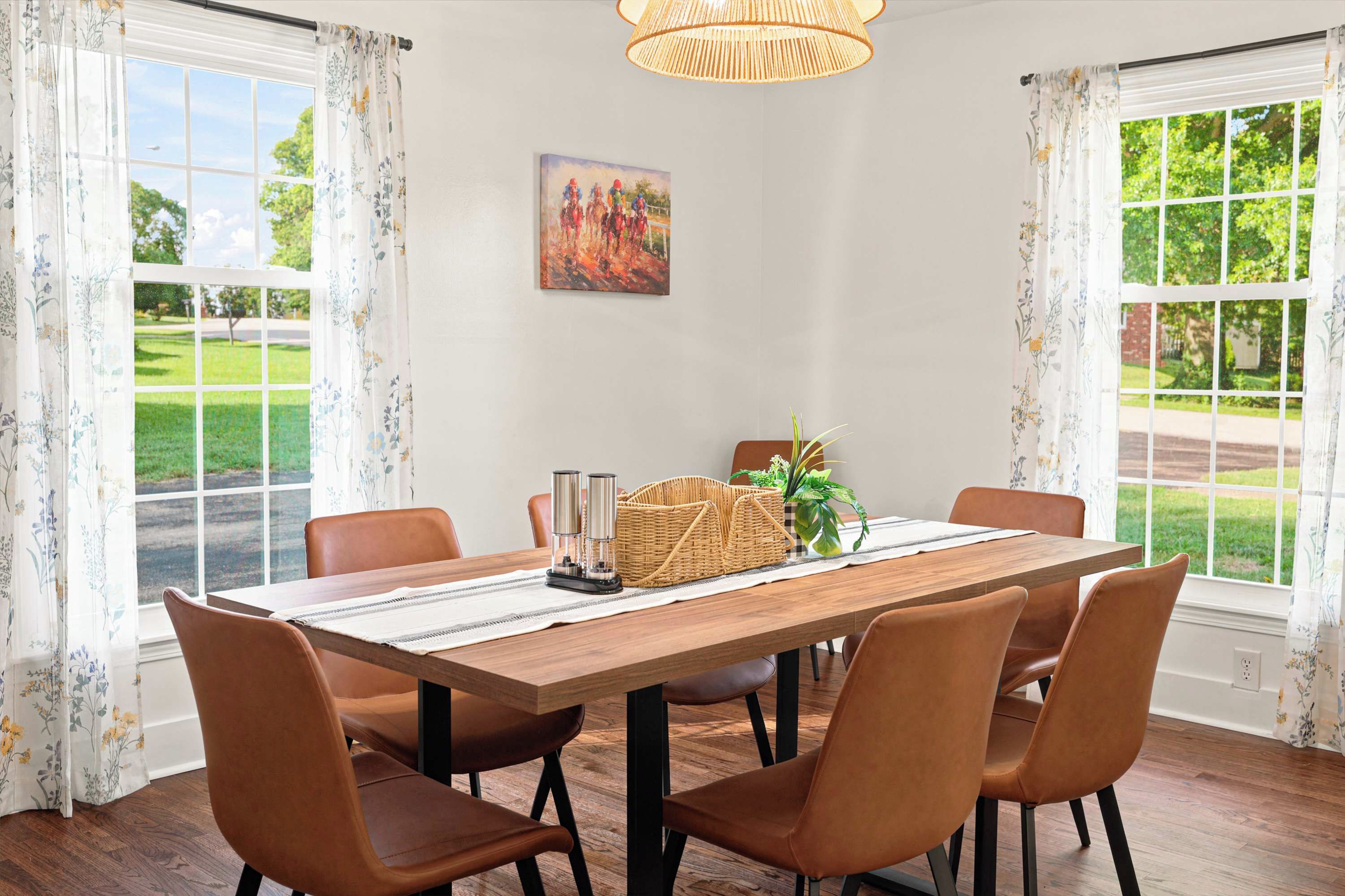 A dining room with a wooden table, four chairs, a painting, and a view of a green landscape through large windows.
