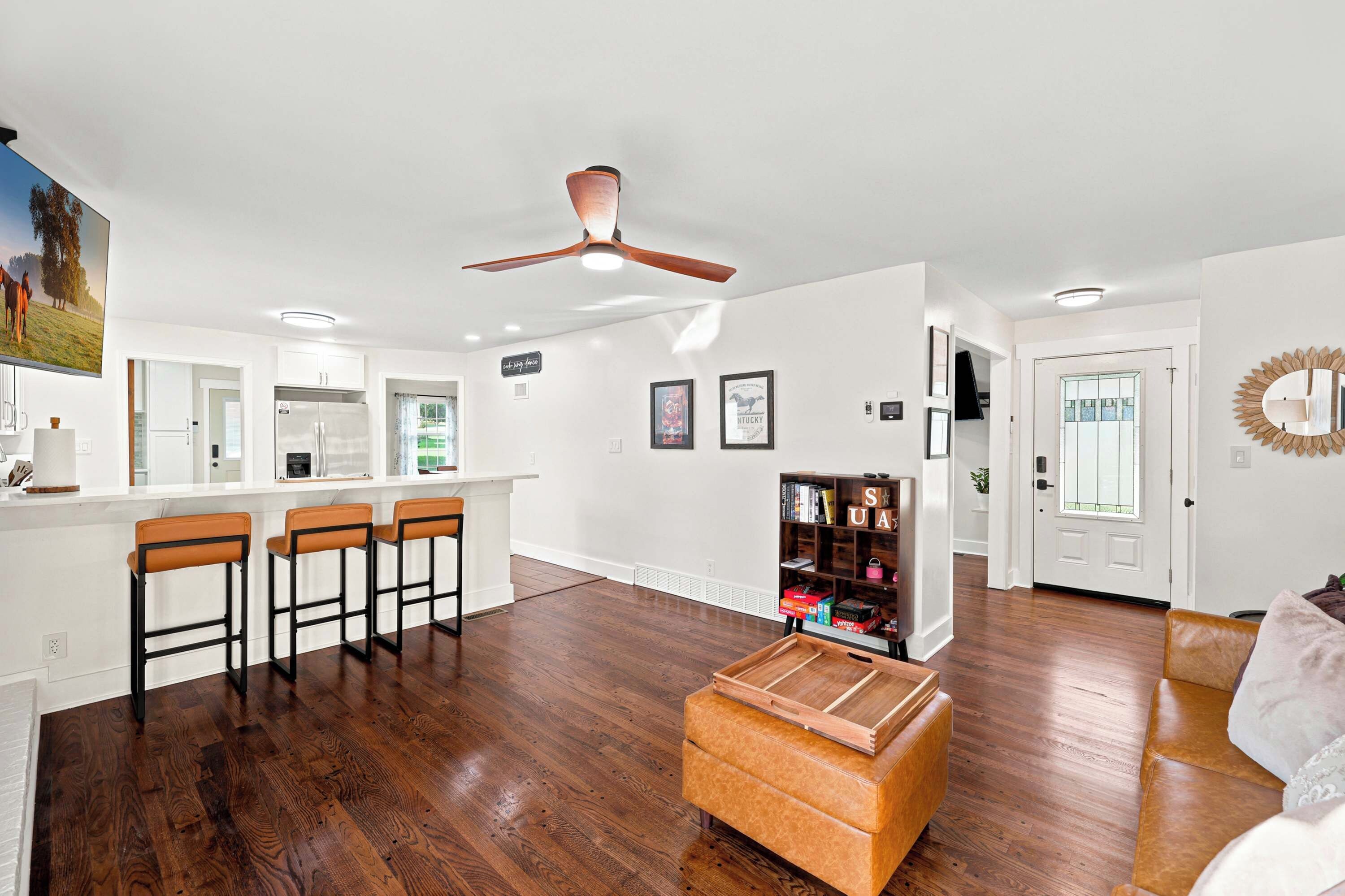 A spacious living area with a wooden floor, a ceiling fan, a bar area with stools, and a bookshelf.