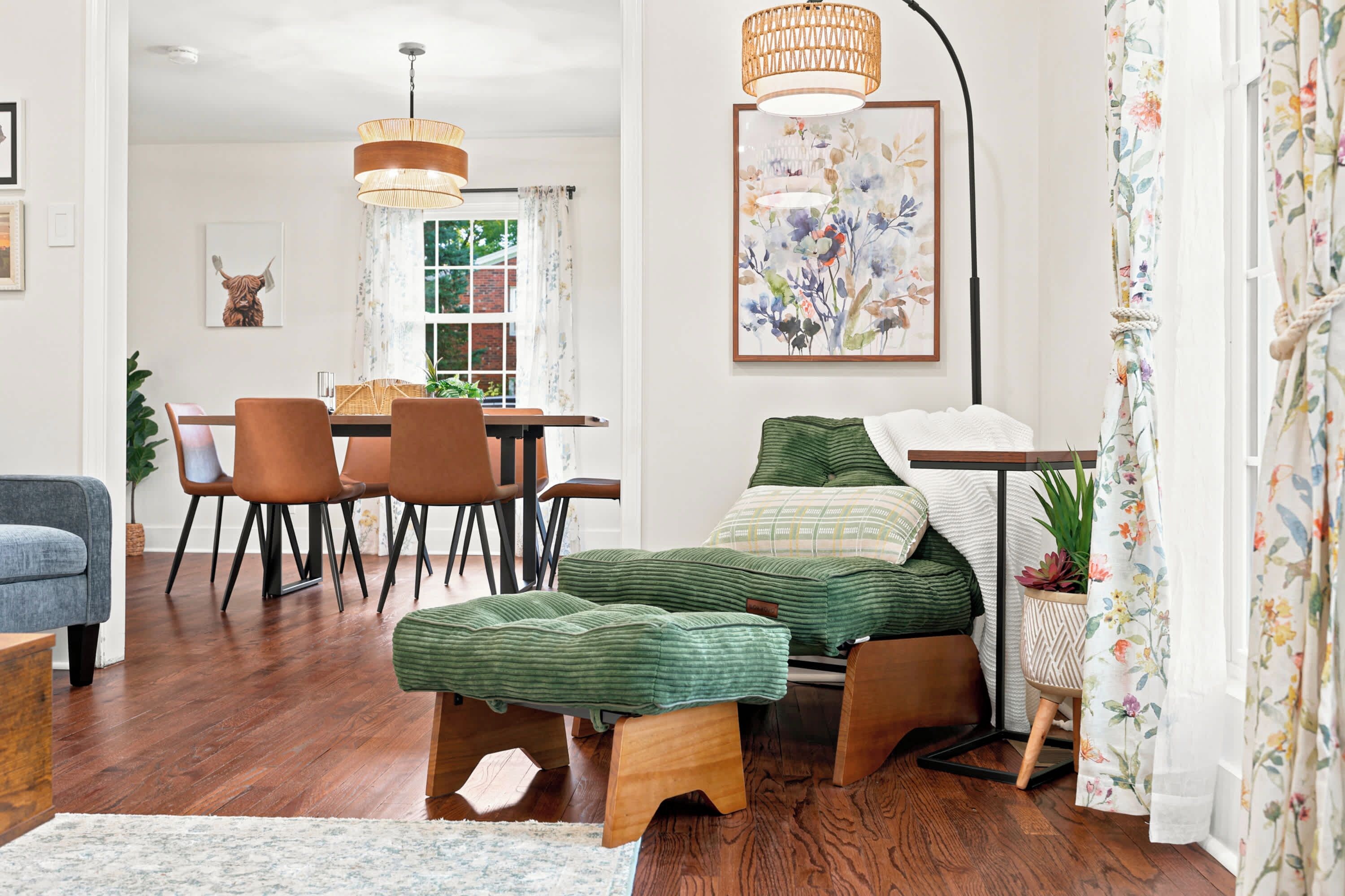 A cozy living room with a sofa, a dining table with chairs, a green pouf, and a wall-mounted lamp.