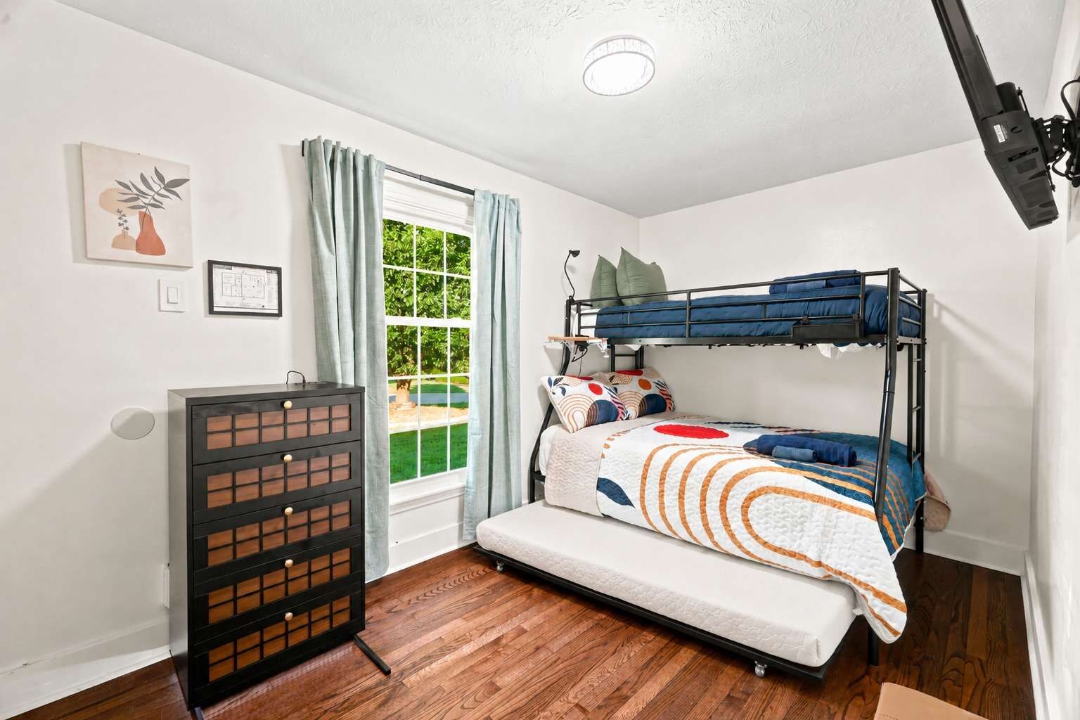 Une petite pièce comprenant un lit superposé, une commode, une fenêtre avec des rideaux et un luminaire au plafond.