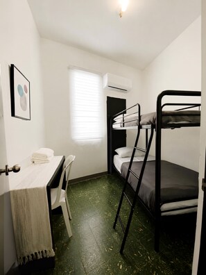 Ein kleines Zimmer mit Etagenbetten, einem Schreibtisch mit Stuhl, einem Fenster mit Jalousien und einer Klimaanlage.
