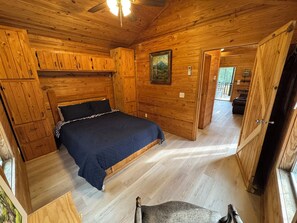 Ein Schlafzimmer einer Holzhütte mit einem Bett, einem Gemälde und einer Tür zu einem anderen Raum.