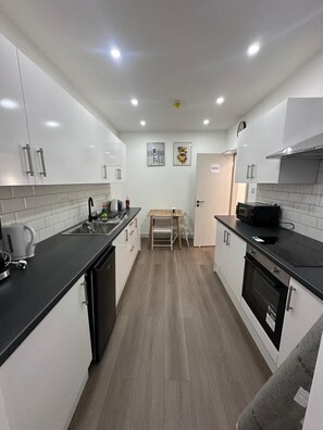 A modern kitchen with white cabinets, a black countertop, and a wooden dining table. - Private Bedroom With Kitchen Access (London)