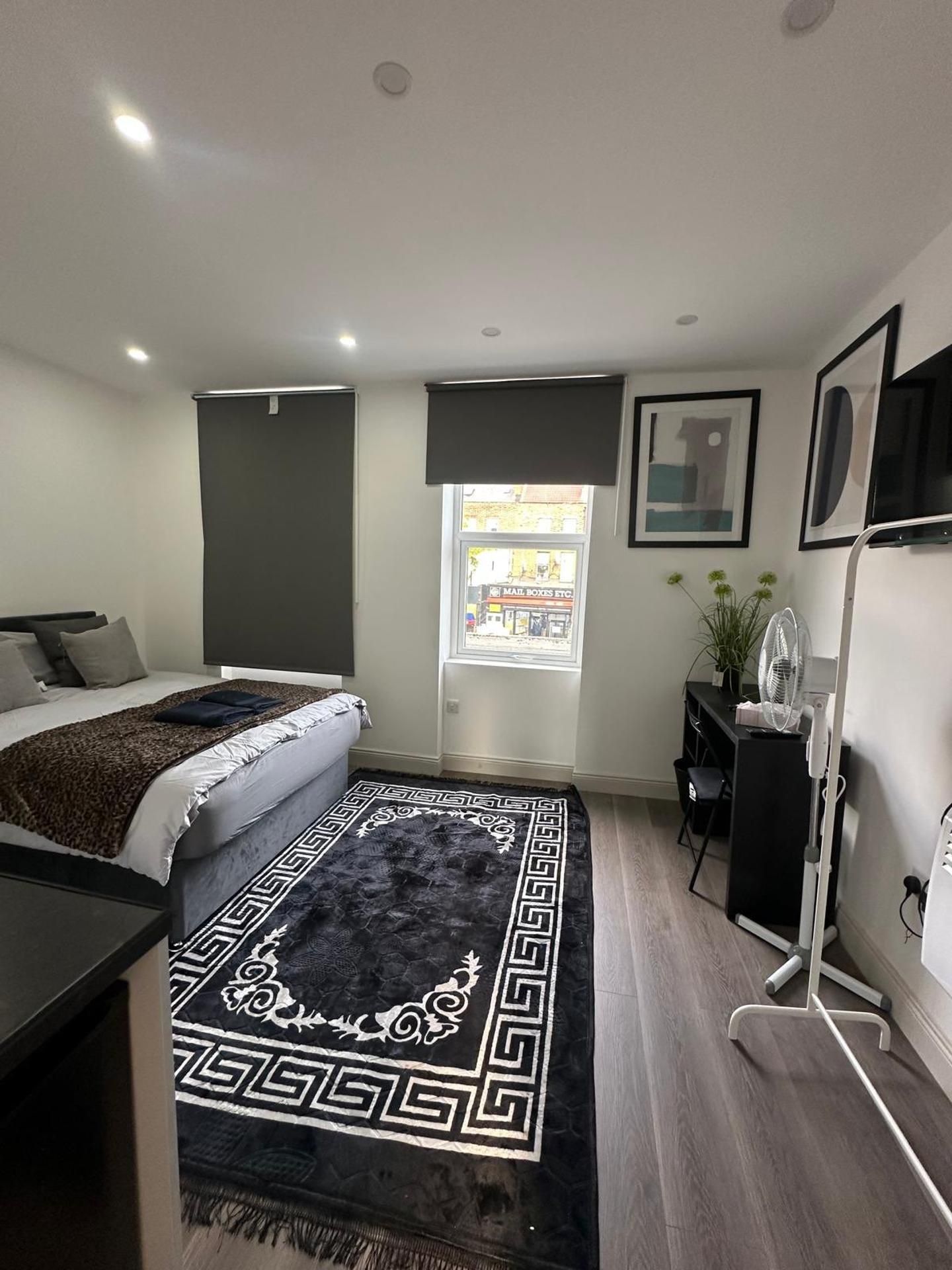 A modern bedroom with a bed, a desk, a chair, a standing fan, and a window with a view of the outside.