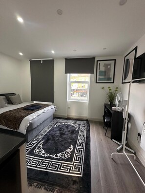 A modern bedroom with a bed, a desk, a chair, a standing fan, and a window with a view of the outside. - Private Bedroom With Kitchen Access (London)