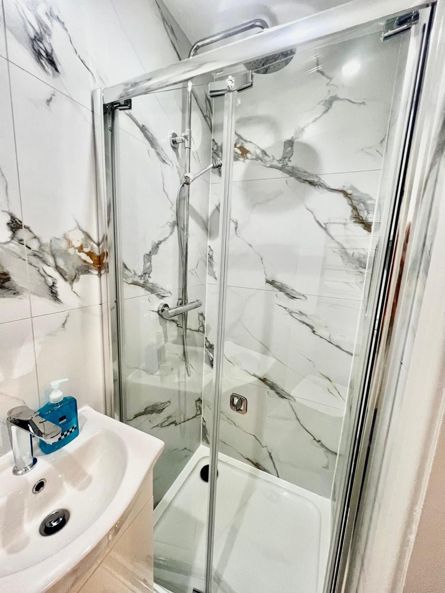A bathroom with a glass-enclosed shower, a marble-tiled wall, and a white sink with a soap dispenser.