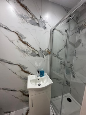 A bathroom with a glass shower enclosure, a white sink with a blue soap dispenser, and marble-patterned walls. - Private Bedroom With Kitchen Access (London)