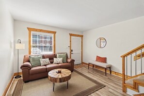 A living room with a brown leather sofa, a round wooden coffee table, a floor lamp, and a window with blinds. - Cozy King Condo Walk to Bojangles & Ovens (Charlotte)