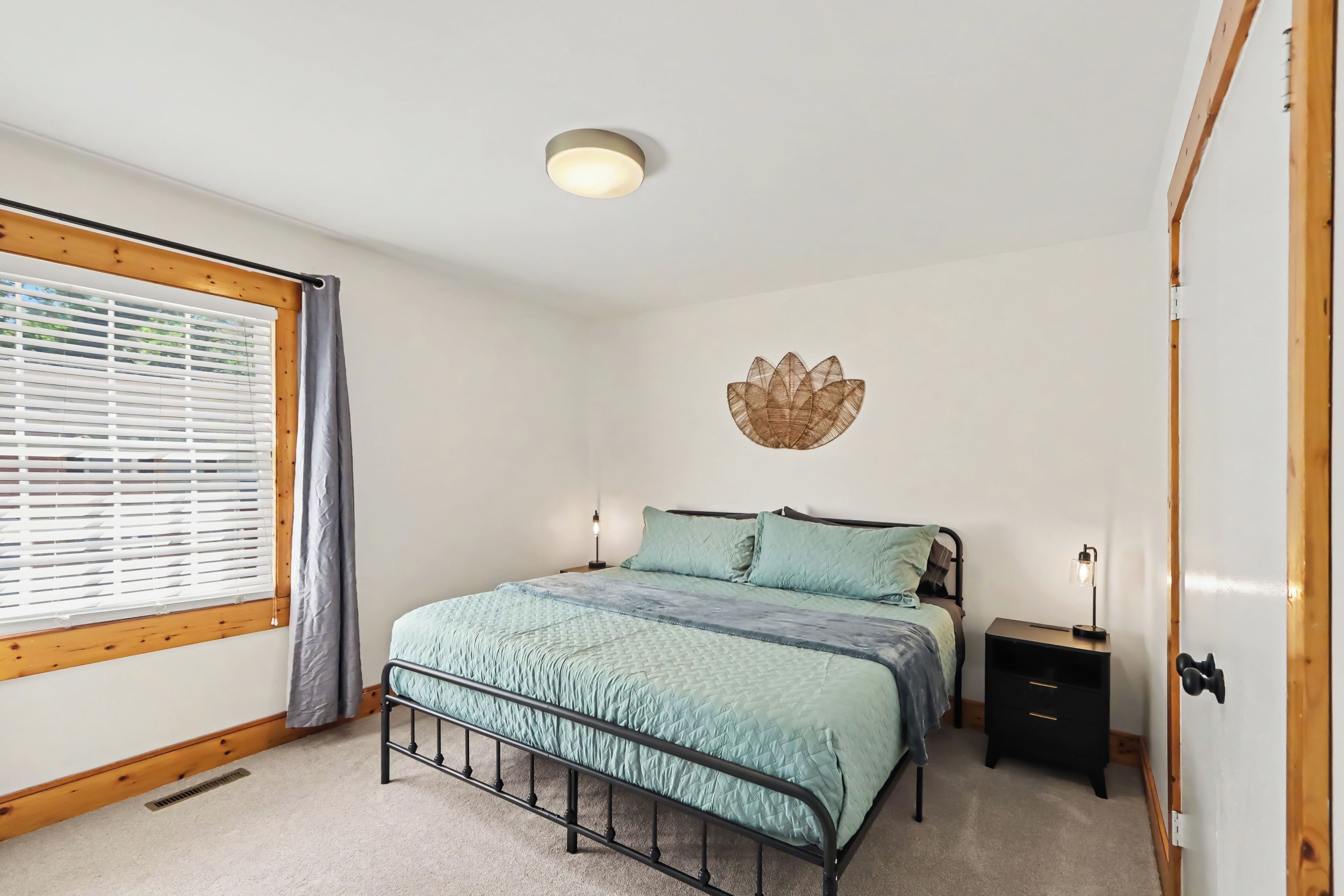 A bedroom with a bed, two bedside tables, a wall-mounted light fixture, a window with blinds, and a door.