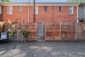 A brick house with a wooden fence, a blue door, and a small garden with a chair and potted plants. - Cozy King Condo Walk to Bojangles & Ovens (Charlotte)